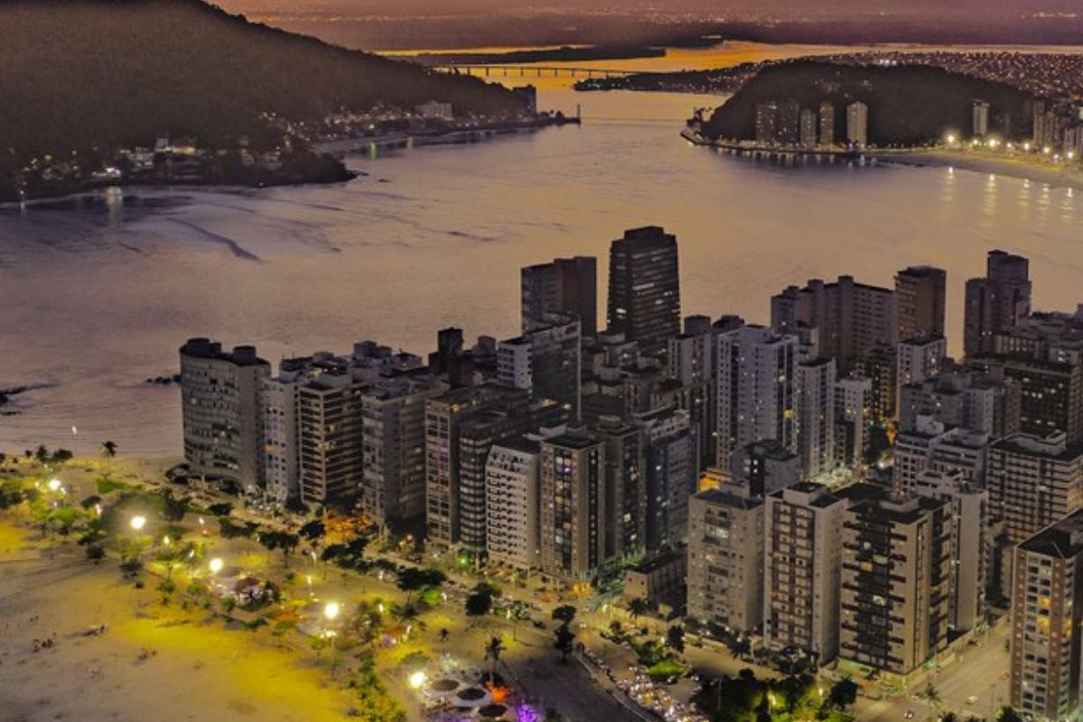 Vista de cima da Estância Balneária em um fim de tarde. Há um aglomerado de grandes prédios, em frente a orla da praia, e atrás o mar