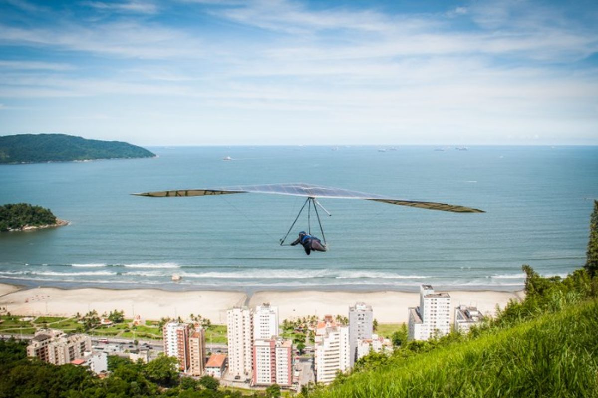 Uma pessoa no ar de asa delta. Ao fundo a cidade de São Vicente, seguida do mar azul