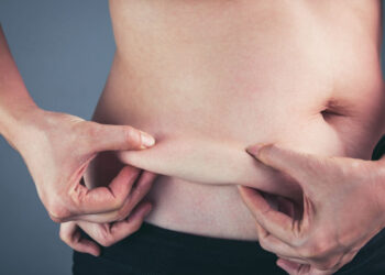Close up female checking fat on abdomen, grey background. Woman's body with flabby stomach holding the excessive belly fat in her hands. Obesity and Overweight Concept. Vintage film filter effect.