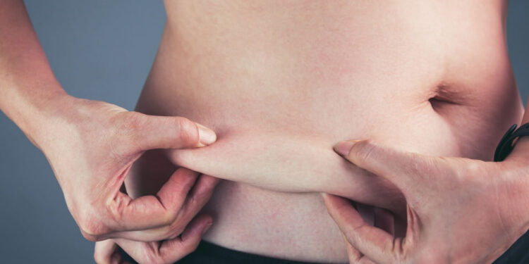 Close up female checking fat on abdomen, grey background. Woman's body with flabby stomach holding the excessive belly fat in her hands. Obesity and Overweight Concept. Vintage film filter effect.
