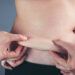 Close up female checking fat on abdomen, grey background. Woman's body with flabby stomach holding the excessive belly fat in her hands. Obesity and Overweight Concept. Vintage film filter effect.